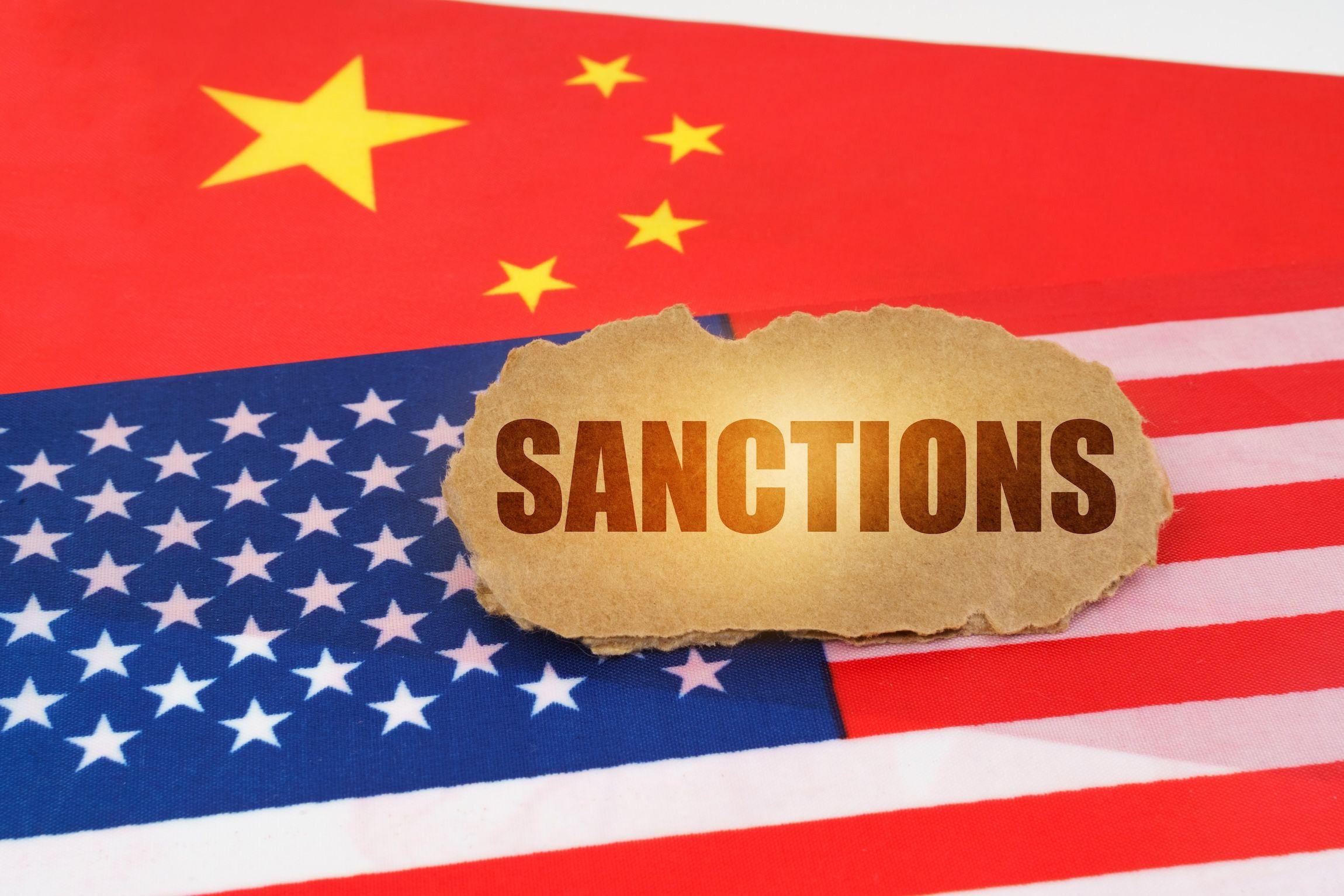 China Politik, China Wirtschaft: die Flaggen von China und USA liegen unter einem Schild mit der Aufschrift "Sanctions" / Sanktionen